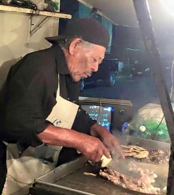 Older man cooking meat on a griddle at a street food stand while wearing a backward cap.