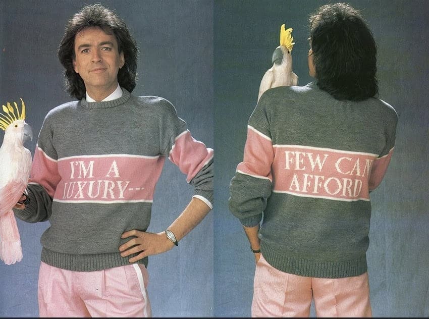 A man with voluminous hair wears a sweater that says “I’m a luxury—” on the front and “Few can afford” on the back while holding a cockatoo.