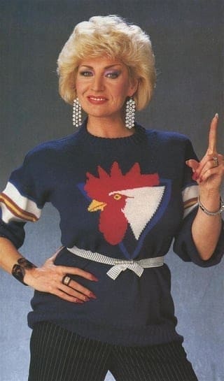 A woman in a dark sweater with a giant rooster graphic, large earrings, and pointing a finger upward.