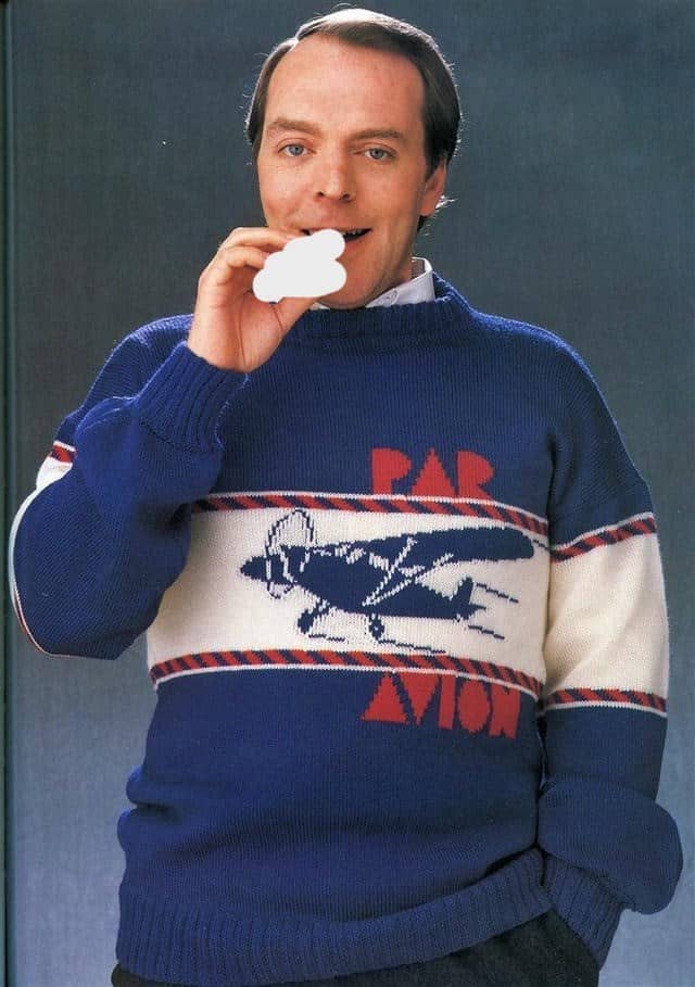 A man in a blue sweater with a vintage airplane graphic and the words “PAR AVION,” holding a cutout of a cloud.