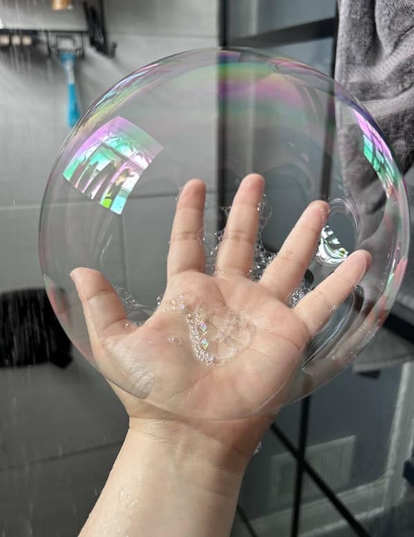 Satisfying photo of a hand holding a giant, nearly perfect soap bubble that glimmers with rainbow reflections and transparency.