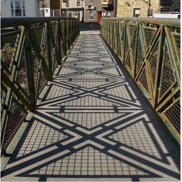 Satisfying photo of a bridge railing creating seamless, geometric crisscross shadows across the walkway, forming an intricate and soothing pattern.