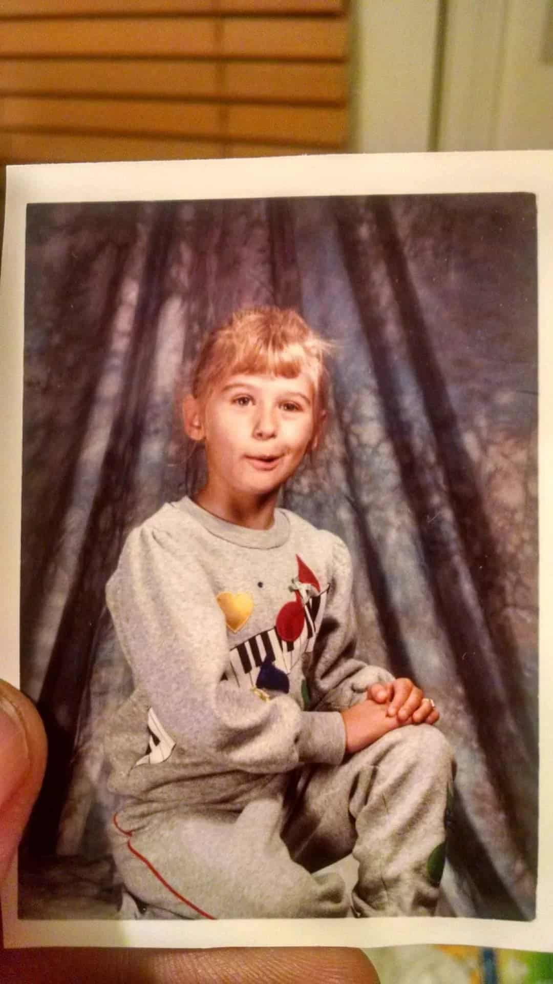 School portrait of a child in a gray sweatsuit with colorful keyboard graphics, bangs, hands clasped, surprised expression