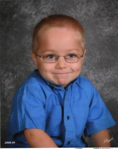 2008–09 school photo of a small boy in glasses and a blue shirt, arms folded with a shy, knowing smile.