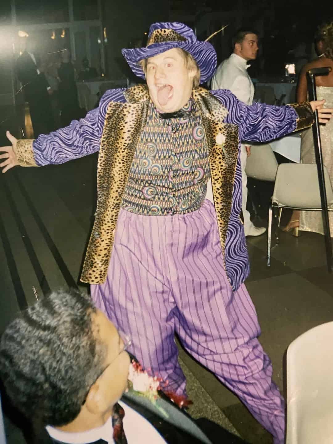 Prom-night snapshot of a teen in a purple hat and leopard-print suit with striped pants, arms wide mid-yell.