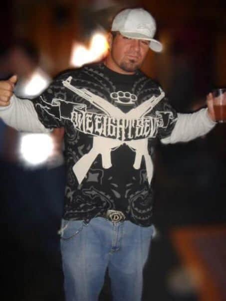 Bar snapshot of a guy in a white cap and layered tees, graphic shirt with crossed rifles, chain belt, drink in hand.