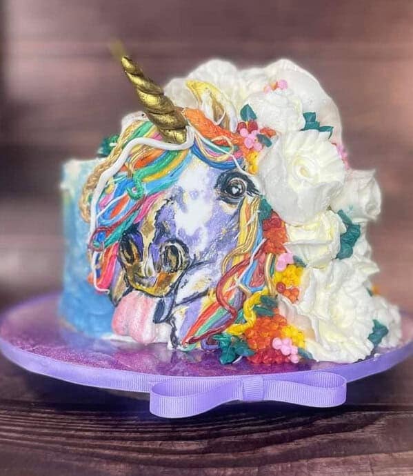 A unicorn cake with a melted, rainbow-colored face and tongue sticking out, surrounded by chaotic frosting flowers.