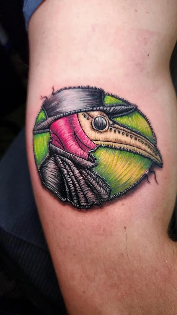 Colorful plague doctor head rendered in stitch-like shading inside a round patch-style tattoo.