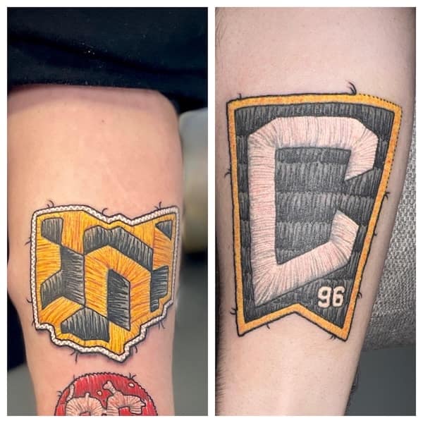 Side-by-side embroidered patch tattoos of sports-style crests in gold and gray with faux thread and frayed edges.