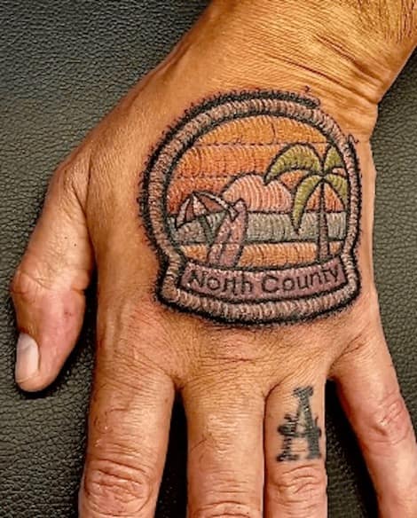 Round beach-badge tattoo on a hand reading “North County” with sunset stripes, surfboards and a palm in stitch style.