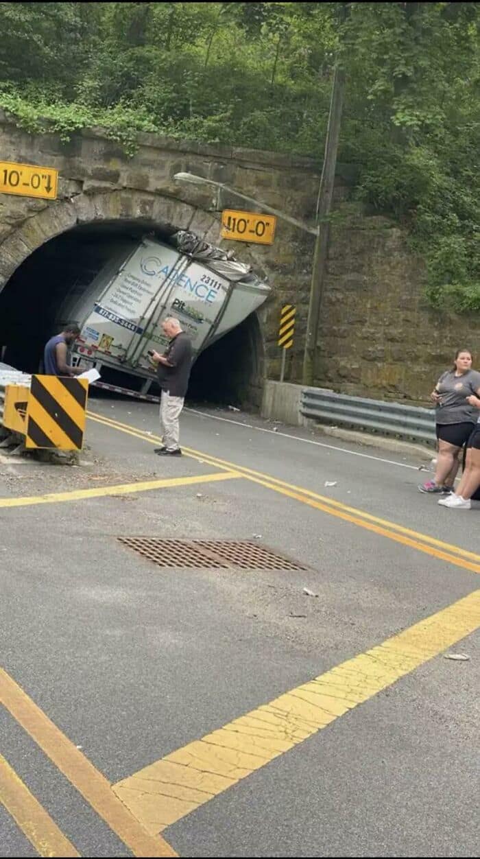 The top of a large semi-truck trailer wedged and crumpled inside a low-clearance tunnel.
