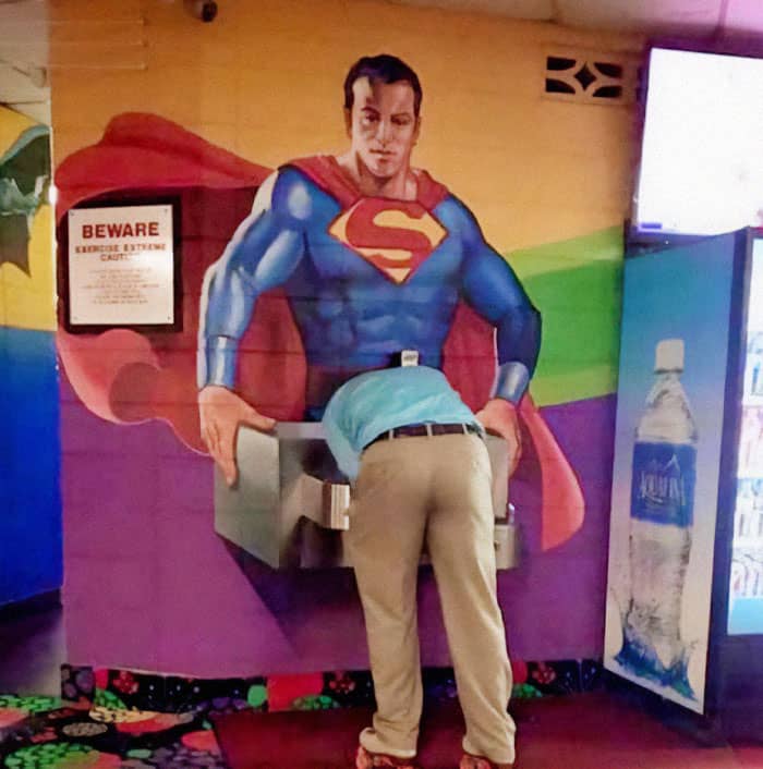 A water fountain placed in a very awkward position on a wall mural of Superman.