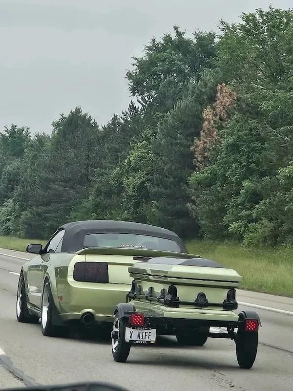 A funny meme of a green Mustang pulling a matching green casket on a trailer with an "X WIFE" license plate.