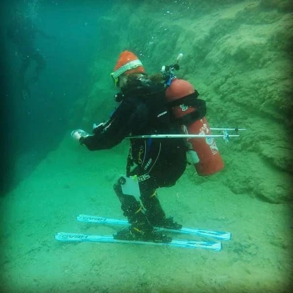 A person in full scuba gear bizarrely wearing skis and a Santa hat underwater.