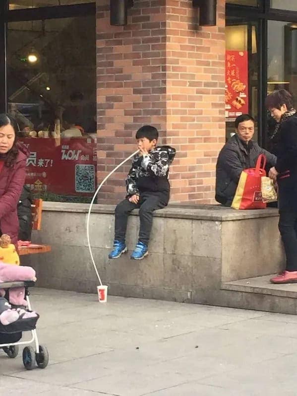 A funny pic of a young boy drinking from a cup on the ground with a very long straw.