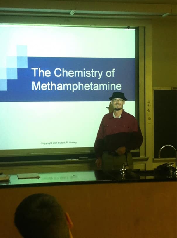 Chemistry teacher in hat standing by slide titled “The Chemistry of Methamphetamine.”