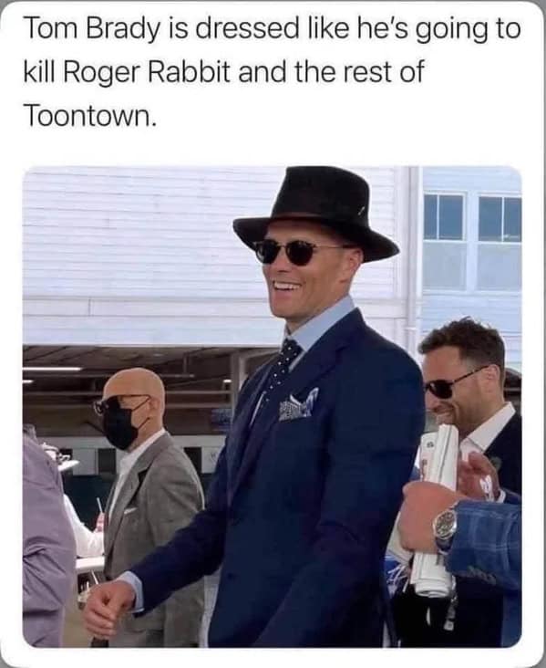 Tom Brady in a navy suit, fedora, and sunglasses smiles while walking at an event.