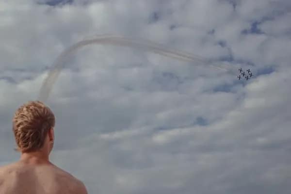 Man on beach watches formation jets draw a curved smoke arc across a cloudy sky.