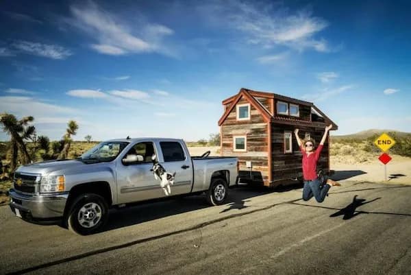 Pickup towing a tiny house; dog hangs out window as owner jumps midair on desert road