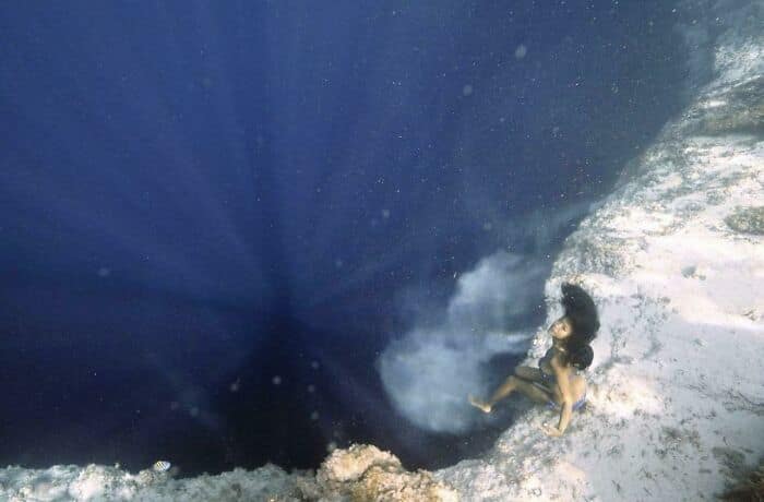 A diver sits on a ledge, dangling her feet over a deep and dark underwater abyss.