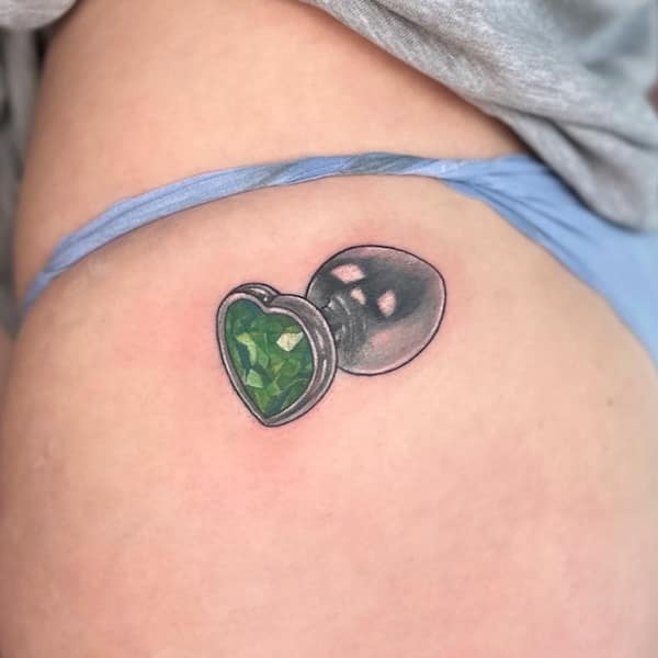 A tattoo of a green gemstone butt plug placed near the hip.