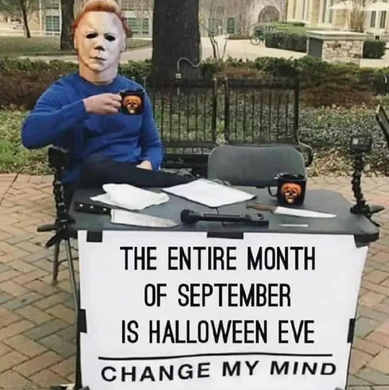 Masked figure sitting at a “Change My Mind” table with a mug; sign reads “The entire month of September is Halloween Eve.”