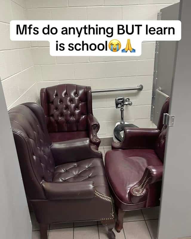 A strange and baffling photo of a high school bathroom stall where the toilet has been removed and bizarrely replaced with two large, fancy leather armchairs.