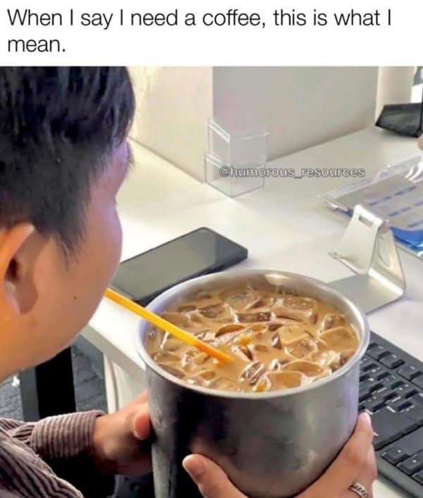 Person at a desk sipping iced coffee from a comically huge metal bucket with a straw near a keyboard and phone.