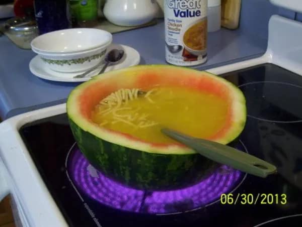 A cursed image of someone cooking chicken noodle soup inside a hollowed-out watermelon on the stove.