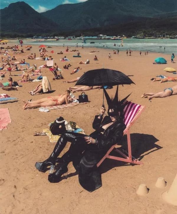 A cursed image of a person in full goth attire, including an umbrella, sitting on a sunny beach.