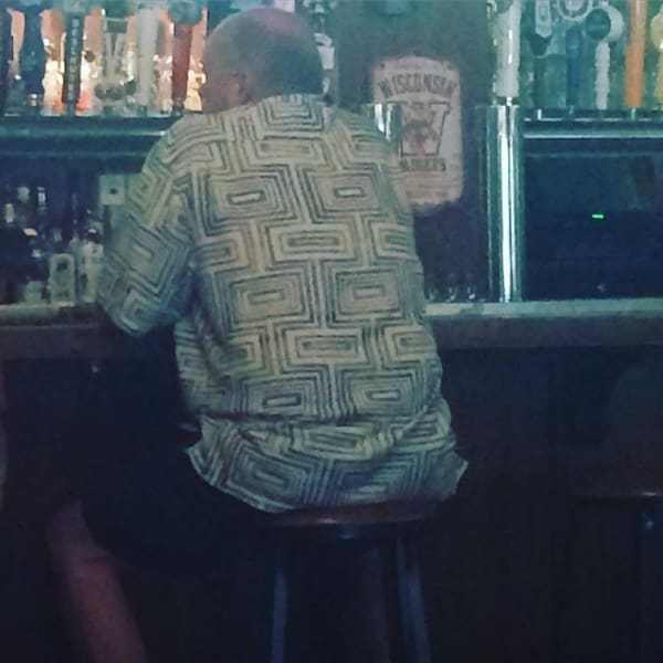 A classic dad fit featuring a loud, patterned, short-sleeve button-up shirt worn while sitting at a bar.