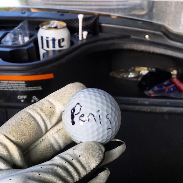 A hilarious found golf ball with the word "Penis" crudely handwritten on it with a black Sharpie marker.