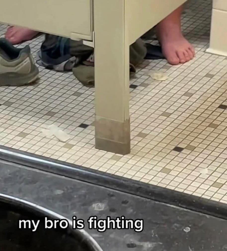 A funny and slightly concerning photo that has been taken in a public bathroom, looking under the stall door, where one of the people inside is barefoot and appears to be fighting.