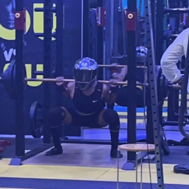 A strange and very funny photo of a person who is in a gym and is doing a very heavy and deep squat while wearing a full, Daft Punk-style motorcycle helmet on their head.