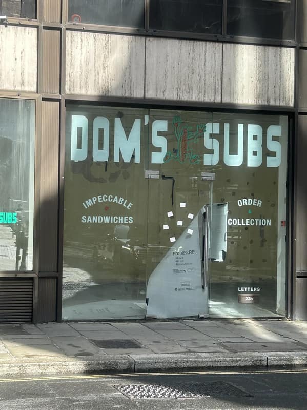 A photo of a sandwich shop called "Dom's Subs" with a large, shattered glass door that looks like a shark bite.