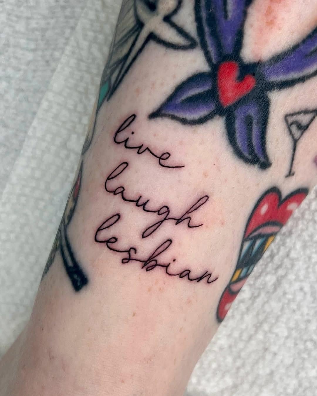 A photo of a funny and creative tattoo on a person's arm that parodies the classic "Live, Laugh, Love" sign, but instead reads "live, laugh, lesbian" in a cursive script.