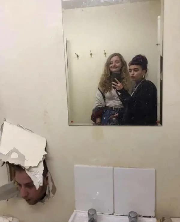 A bizarre and chaotic photo of two women who are taking a selfie in a dirty bathroom mirror, while a man's head is inexplicably sticking through a large glory hole in the wall below them.
