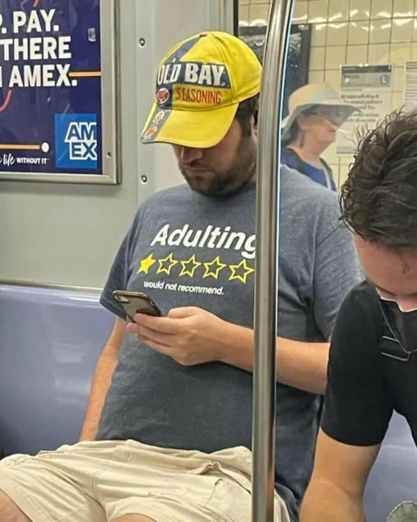 A candid photo of a man on a subway who is wearing an "Old Bay Seasoning" hat and a t-shirt that gives the concept of "Adulting" a one-star review.