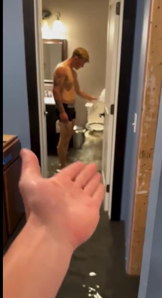 A hilarious first-person photo of a completely flooded bathroom, with a hand gesturing in defeat towards a man who is standing by the overflowing and spraying toilet.