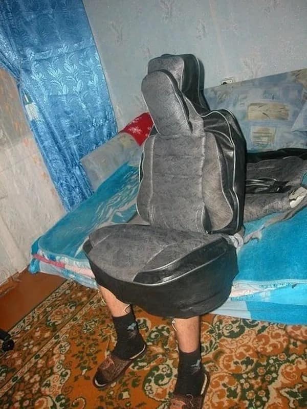 A very surreal and bizarre photo of a person who is sitting on a bed, but their entire torso and their head are completely covered by a gray and black car seat, making them look like a strange creature.