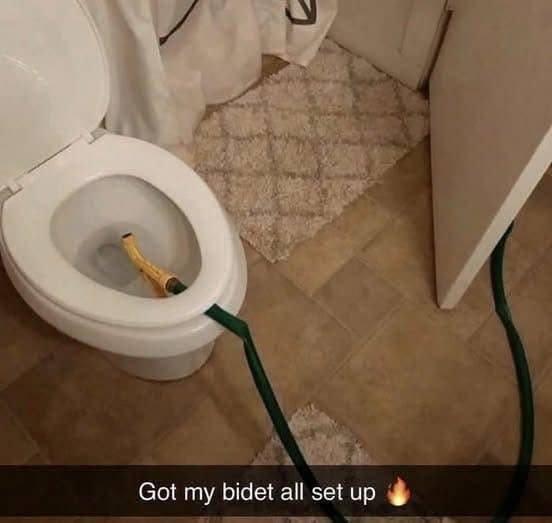 A cursed and hilarious photo of a very questionable DIY bidet that has been constructed in a bathroom by using a green garden hose and a high-pressure spray nozzle.