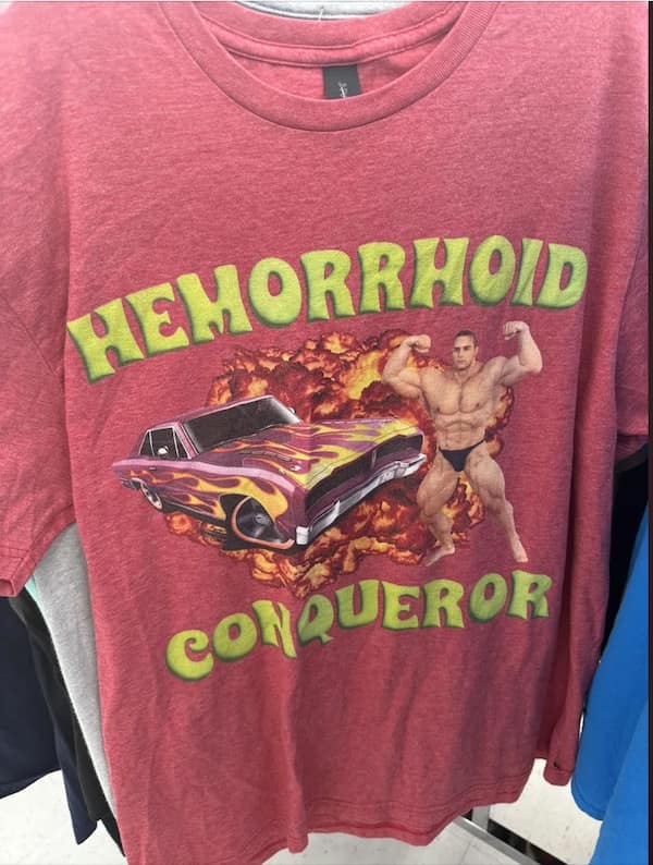 A photo of a truly bizarre and hilarious bright red t-shirt that was found at a thrift store, which has a graphic of a muscle man and a hot rod and says "HEMORRHOID CONQUEROR."