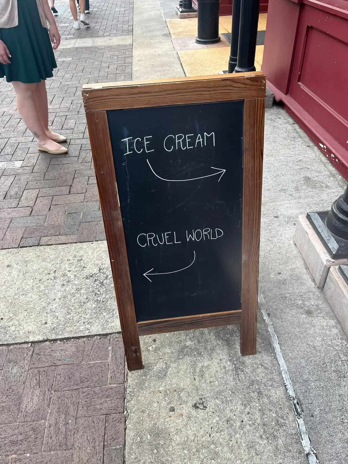 Sidewalk chalkboard with arrows: right to “ICE CREAM,” left to “CRUEL WORLD.”