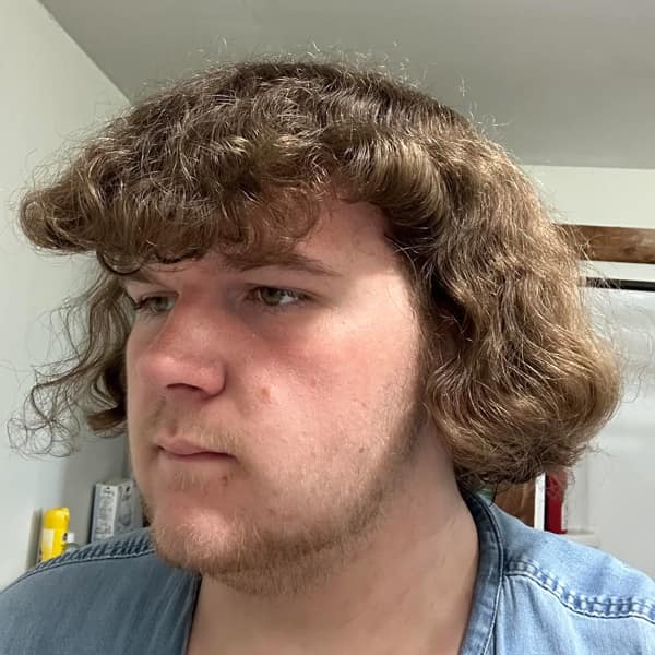 Young man with dense curly, bowl-shaped helmet hair and faint beard, lit by bathroom vanity light.