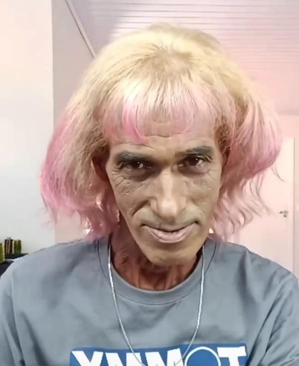 Older man wearing an uneven pink-blond bob with short blunt bangs, smirking in a gray T-shirt indoors.