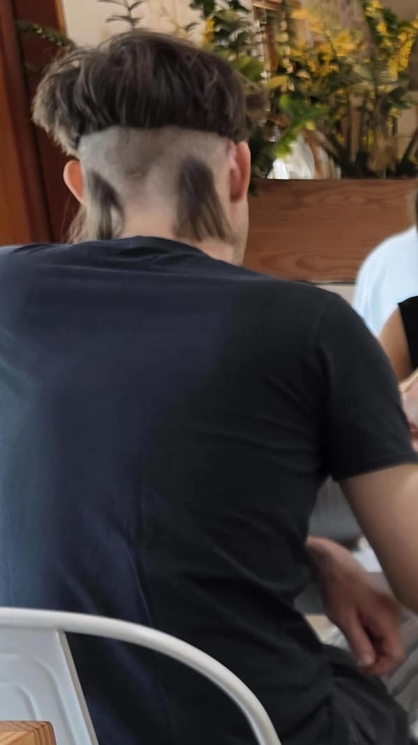 Back view of a man with a bowl-cut ring on top and shaved nape, leaving two dark tufts near the ears.