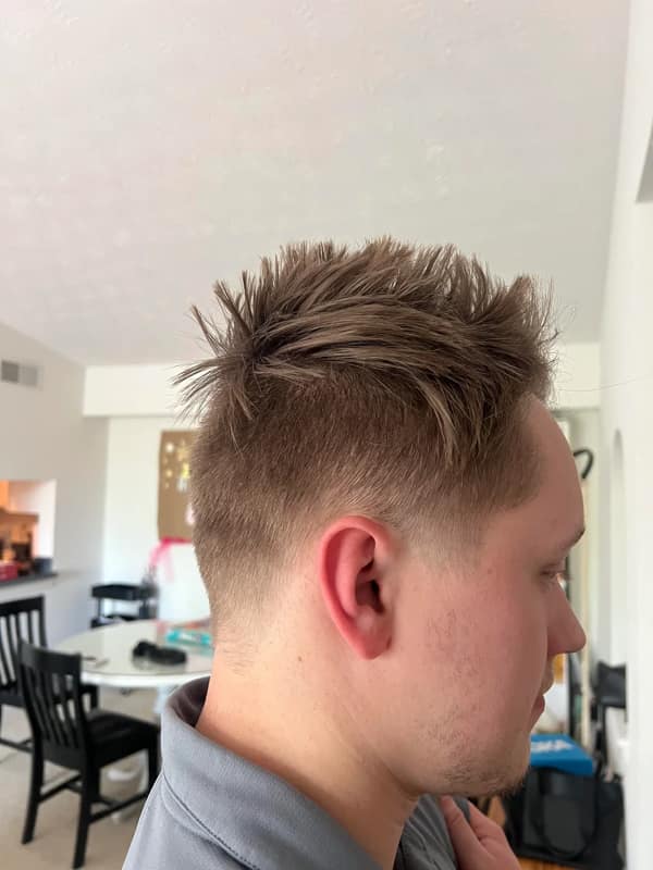 Young man with a high fade and off-center spiky ridge along the crown, indoors by a kitchen table.
