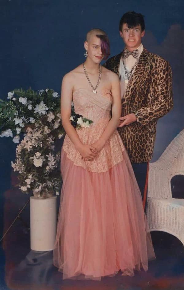 Retro prom photo—teen in pink gown with half-shaved head and purple fringe; date in leopard tux grins.