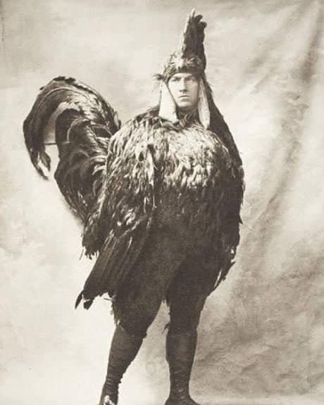A serious-looking man stands proudly while wearing a massive, realistic rooster costume in this old black and white photo.