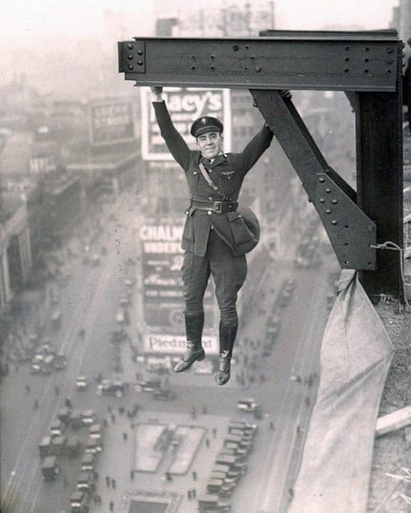 A man in uniform smiles while hanging from a steel girder high above a city in a shocking historical photo.